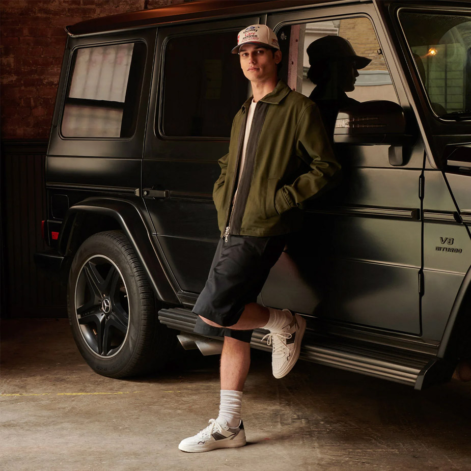 Style shot of male model leaning against a black mercedes with an olive jacket, dark grey shorts, and The Kingston sneakers on.